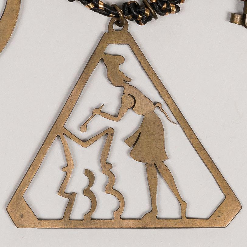 10. Triangular charm with the stylized figure of a female cook stirring a pot The pot forms Greta Perlman s transport number, M 433 and alludes to her work in the camp kitchen.