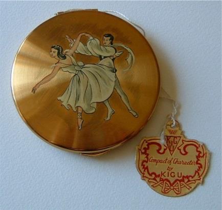 Transfer printed ballet dancers on satin gilt lid. Inner lid with Kigu trademark on a textured surface and a sliding catch. Pink puff with trademark stamped in gold. Framed mirror.