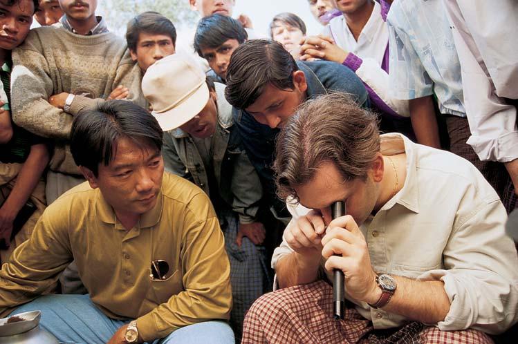 Figure 1. Shown here buying gems in a Mogok, Myanmar (Burma) gem market, the author uses a 10 darkfield loupe to view the interior of a red spinel crystal being offered for sale.