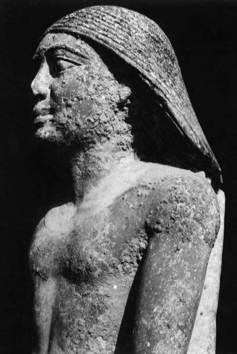 A wig with a center part covers the head of the statue, flaring into triangular panels that end just above the shoulders on either side.