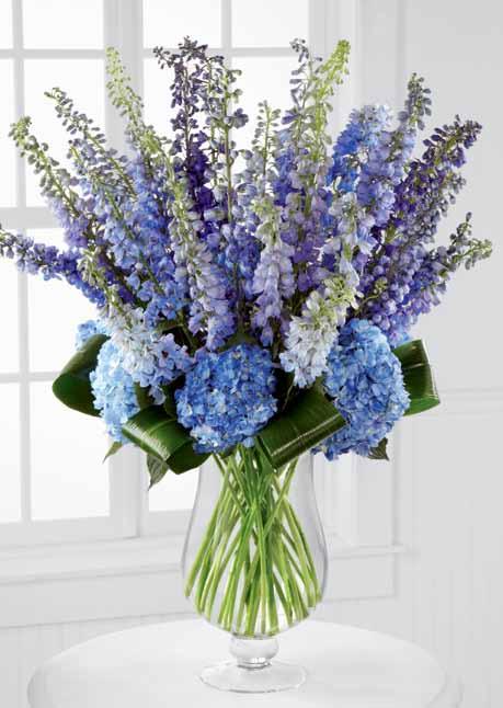 LX117 honestly 12 Blue Hybrid Delphinium stems 12 Purple Hybrid Delphinium stems 7 dark Blue Hydrangea blooms 7 aspidistra leaves 1 Glass Footed Vase (#LUX-Footed) Approx.