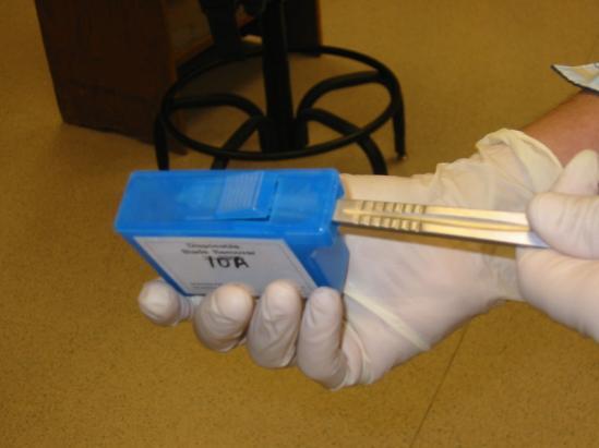 The newer method for blade removal inserts the dull blade into a sharps container as the blade is removed. The blade still on the handle is inserted into the small container as shown below.
