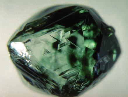 3-30: Right, Strongly resorbed, octahedral diamond with clusters of dark green surface spots and trigons (0.18 ct). Below, Close-up of the dark green spots and trigons. The field of view is ~1.5 mm.